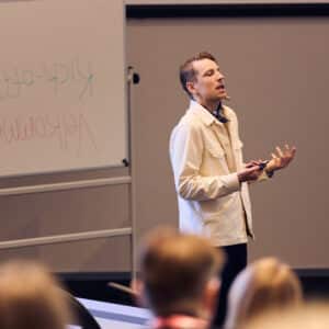 Simon Linde holder oplæg foran en forsamling i et auditorium. Han taler engageret og gestikulerer (på italiensk) med hænderne